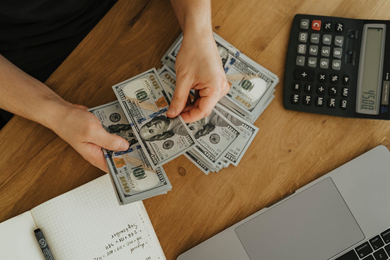 Close-up of hands counting dollar bills with calculator, notebook, and laptop on wooden table.